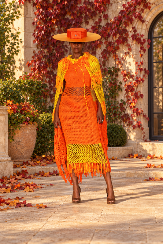 Model wearing the orange and yellow Hand Crochet Fringe Dress and matching Shawl, with a broad-brimmed hat, on a stone terrace with autumn foliage.